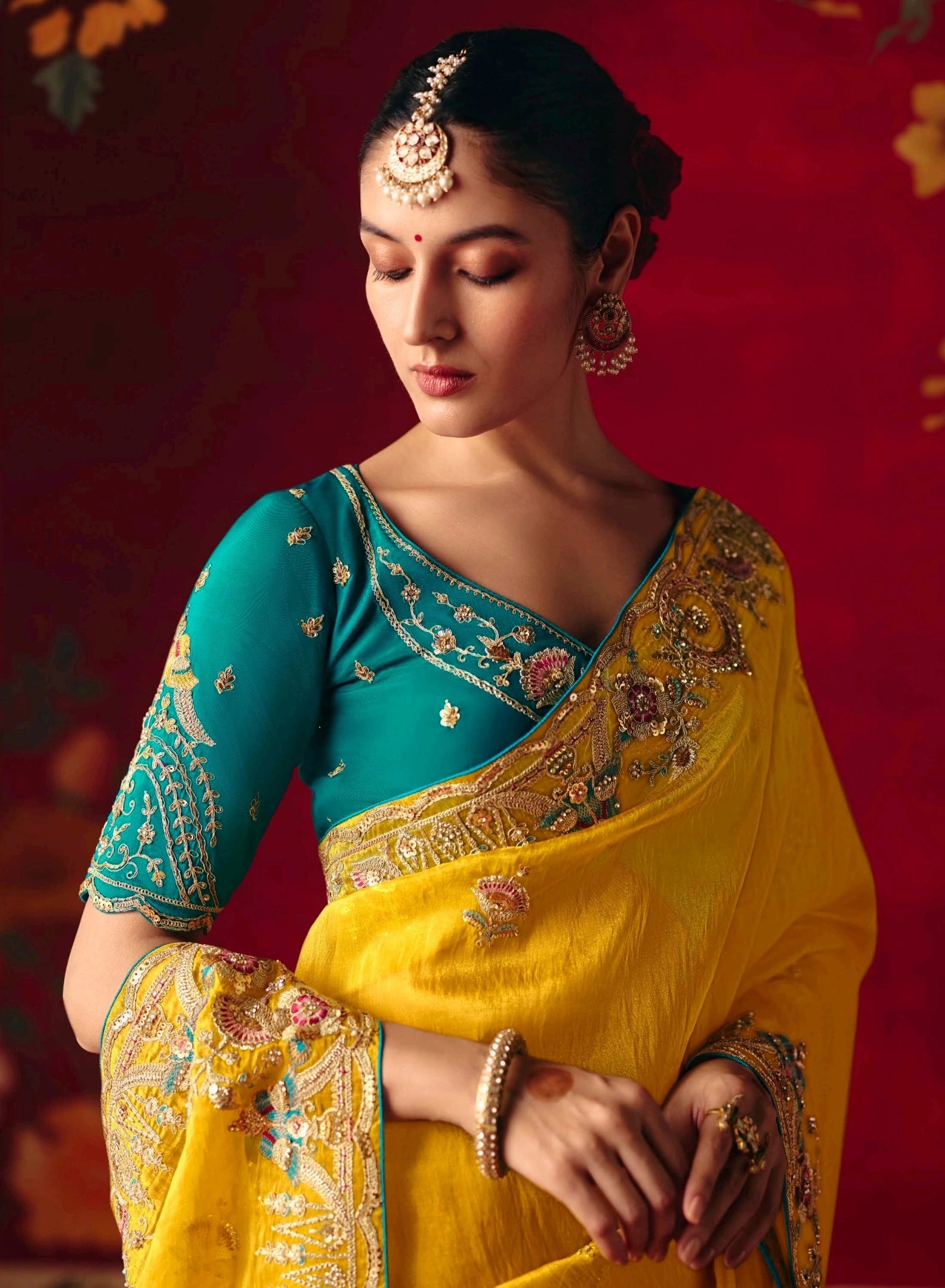 Woman in a yellow and teal saree with intricate patterns against a red background